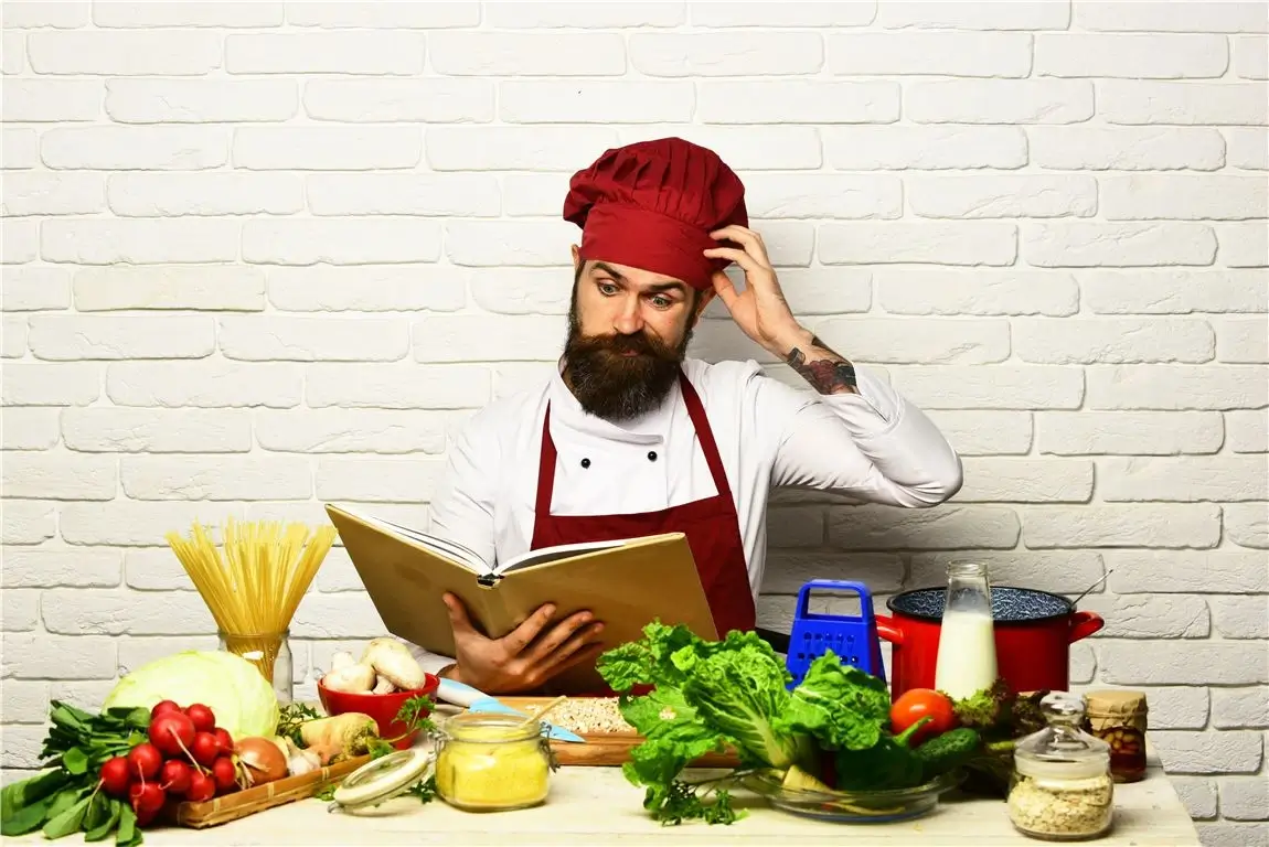 A chef looks puzzled as he reads a cookbook in front of a selection of ingredients